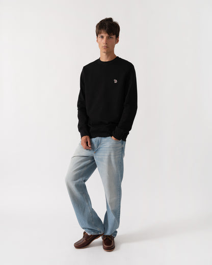A man modeling a black crewneck sweatshirt with a small embroidered logo on the left chest, paired with light blue jeans and brown leather shoes, against a plain white studio background.