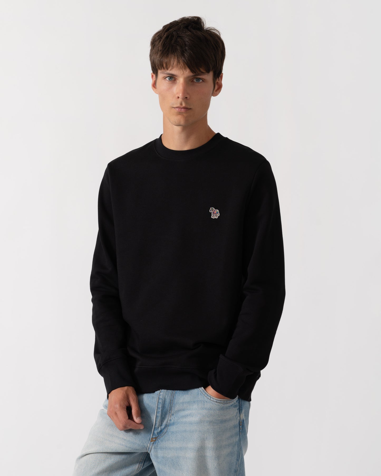 A man wearing a black crewneck sweatshirt with a small embroidered logo on the left chest, paired with light blue jeans, against a plain light gray background.