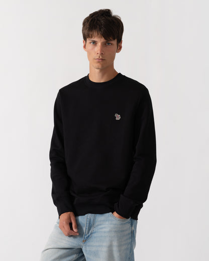 A man wearing a black crewneck sweatshirt with a small embroidered logo on the left chest, paired with light blue jeans, against a plain light gray background.