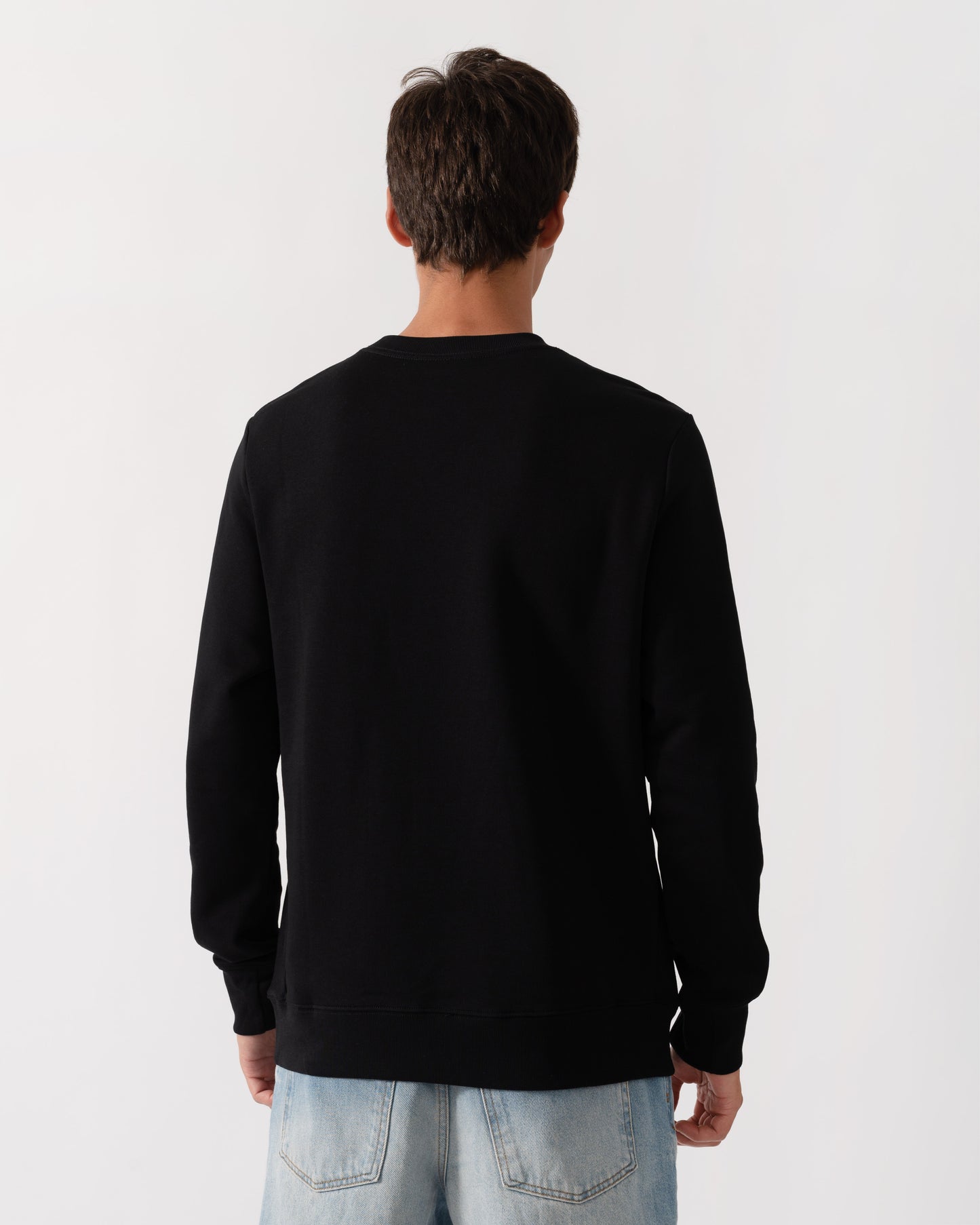 A man viewed from behind wearing a plain black crewneck sweatshirt and light blue jeans, standing against a light gray studio background.