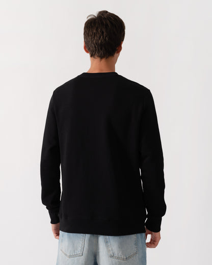 A man viewed from behind wearing a plain black crewneck sweatshirt and light blue jeans, standing against a light gray studio background.
