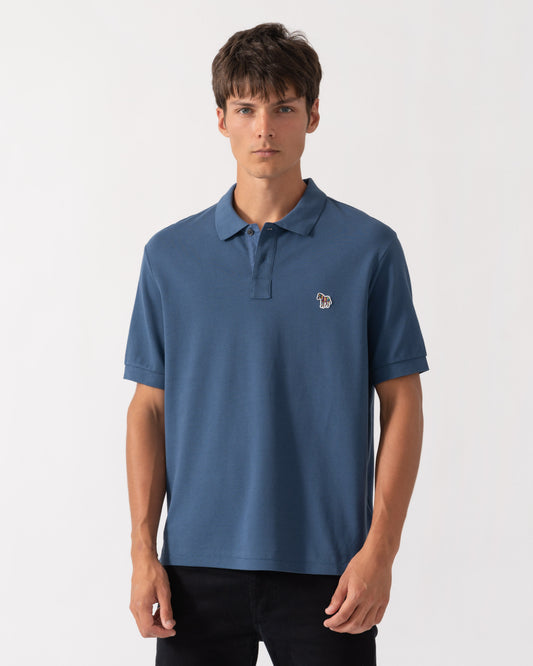 A man wearing a slate blue short-sleeve polo shirt with a small embroidered lion logo on the left chest, paired with black pants, against a plain light gray background.