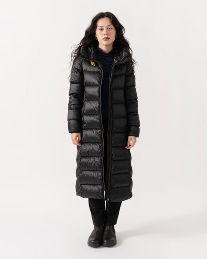 A woman modeling a long black puffer coat with horizontal quilting, a high collar, and a front zipper, photographed against a plain light gray studio background.