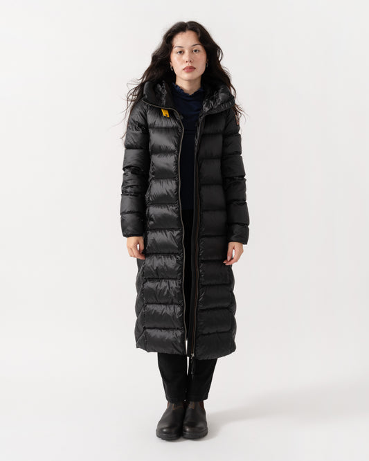 A woman modeling a long black puffer coat with horizontal quilting, a high collar, and a front zipper, photographed against a plain light gray studio background.
