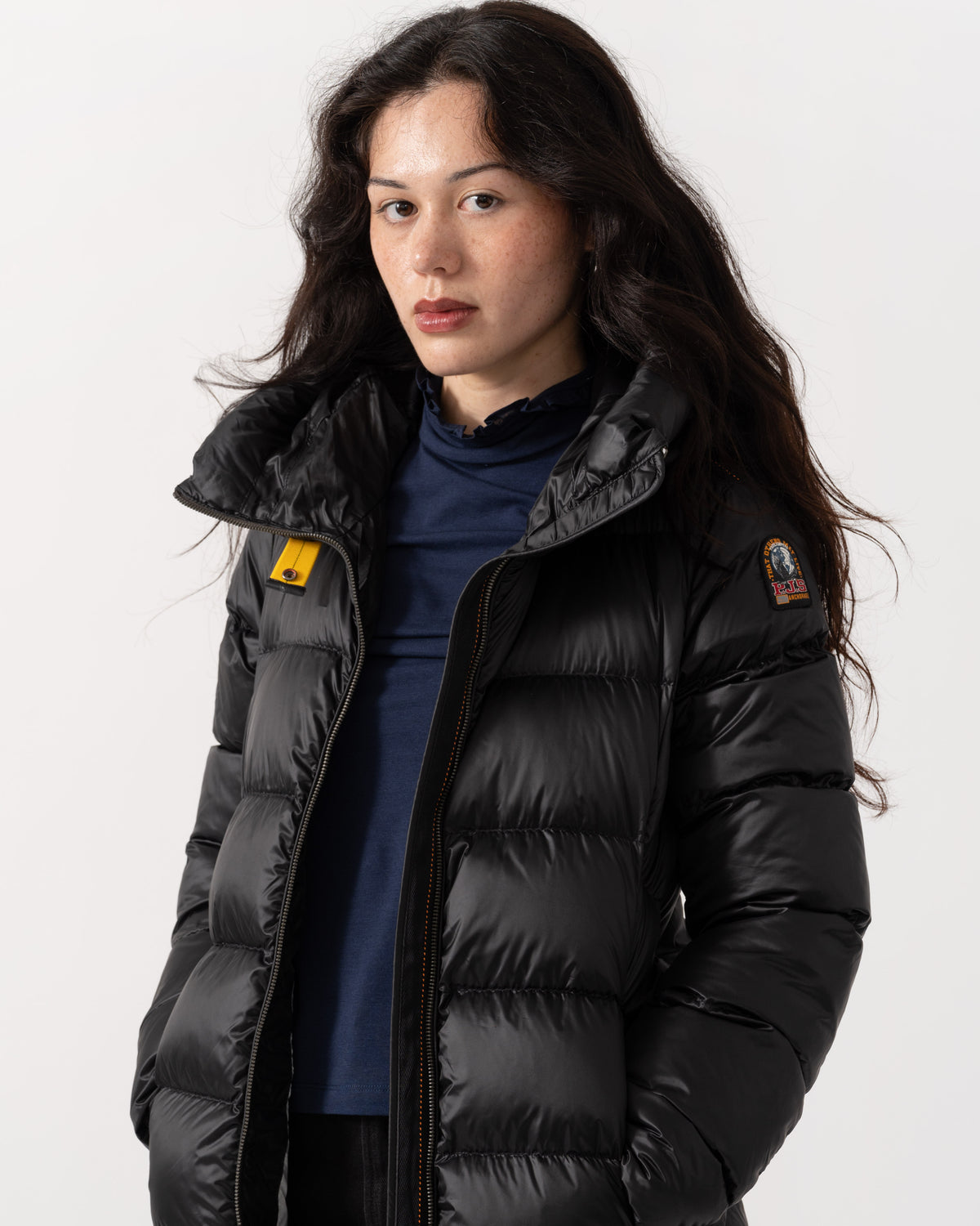 A woman wearing a black puffer jacket with a high collar over a navy blue top, photographed against a clean white studio background.