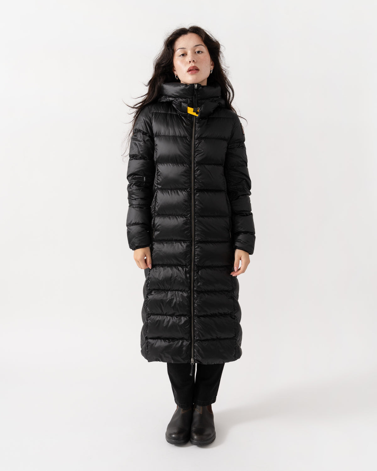 A woman modeling a long black puffer coat with a high collar and a small yellow accent near the zipper, standing against a plain white studio background.