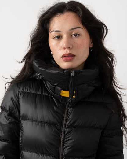 A woman with long dark hair wearing a black puffer jacket with a high collar and a small yellow accent near the zipper, photographed against a plain light background.