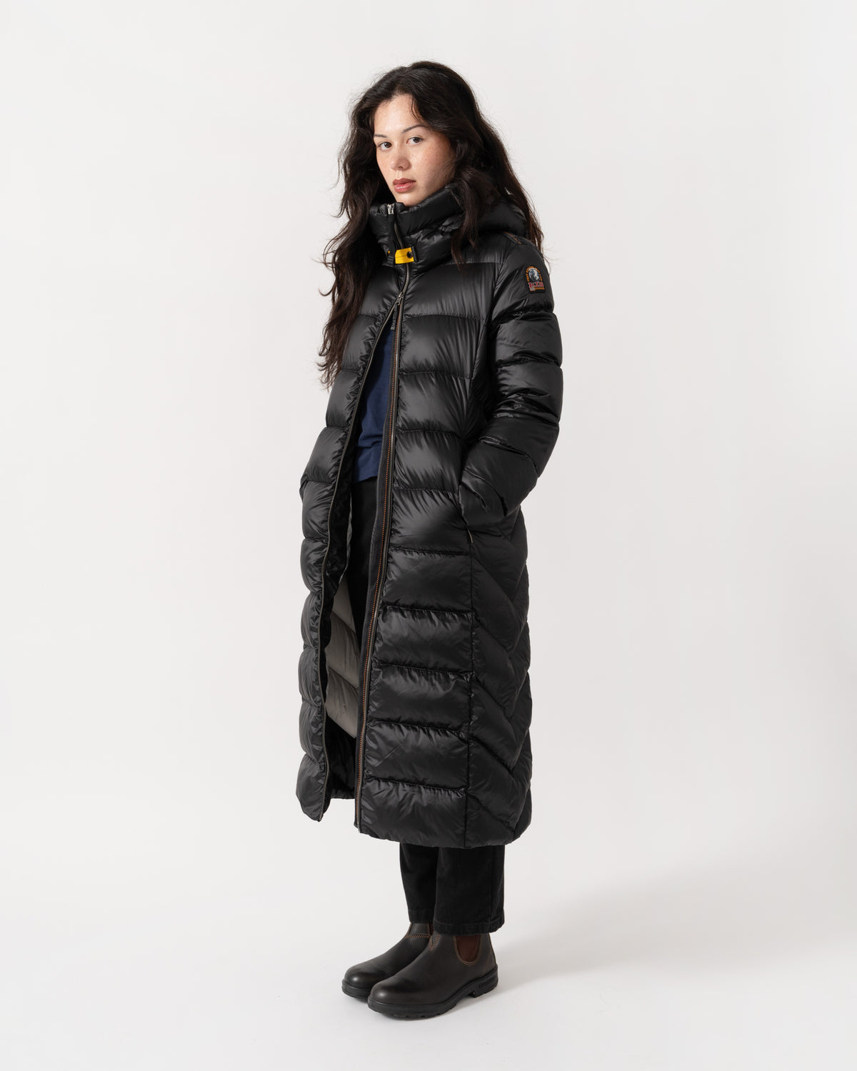 A woman modeling a long black puffer coat with a high collar and horizontal quilting, standing against a plain light gray studio background.