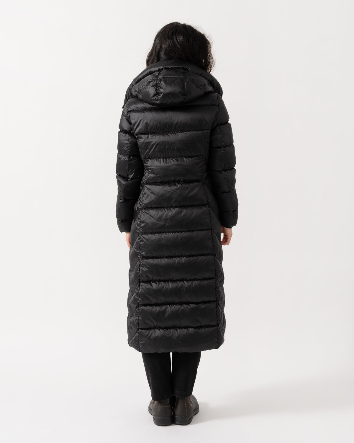 A person viewed from the back wearing a long black puffer coat with horizontal quilting, black leggings, and dark boots against a plain light gray studio background.