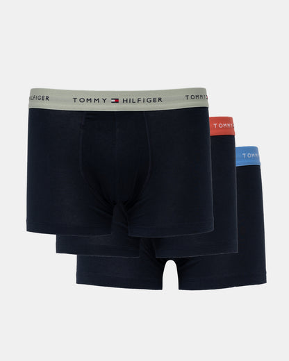 Three pairs of Tommy Hilfiger men's boxer briefs in dark navy blue, stacked together on a light gray background. The front pair has a light gray waistband with the Tommy Hilfiger logo, while the pairs behind it have red, orange, and blue waistbands.
