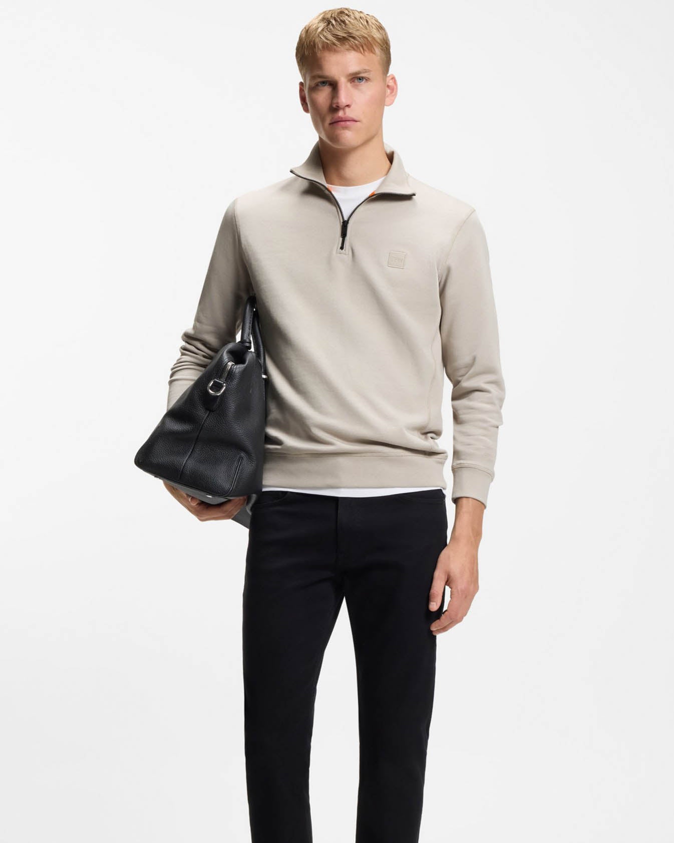 Man wearing a beige sweater and black pants holding a black bag on a white background